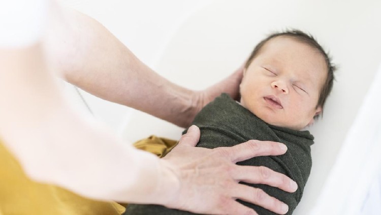new born Mother gently placing her sleeping newborn baby in the bassinet wrapped tightly in a forest green swaddle.