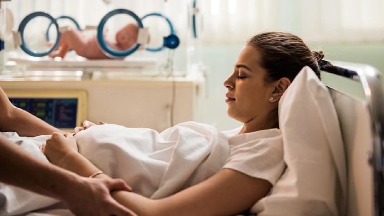 Young woman lying in hospital bed with her eyes closed after childbirth while unrecognizable person is consoling her.