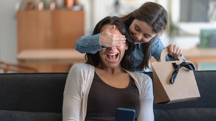 Excited mom being surprised by daughter who is covering her eyes and holding a gift for mother's day - Both smiling
