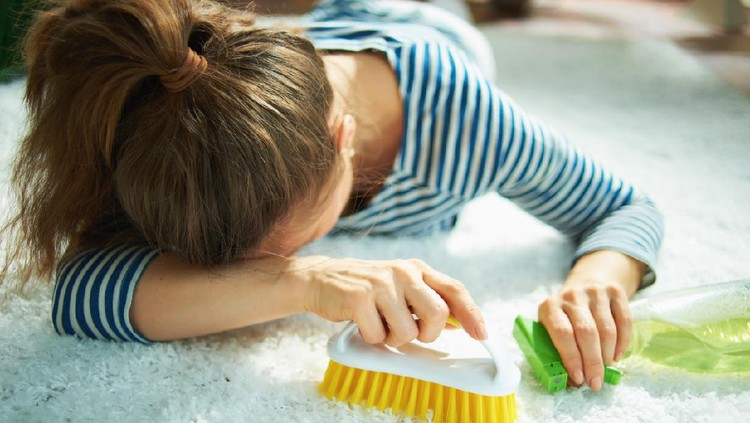 cerita bunda Closeup on tired woman in striped t-shirt and white pants with spray bottle of green cleaning supplies and yellow brush laying on white carpet at home in sunny day.
