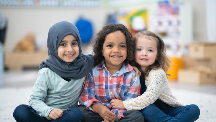 sekolah tk Two girls and a boy are indoors in their preschool classroom. The multi-ethnic group is smiling at the camera while sitting on the carpet and embracing. One girl is Muslim and she is wearing a hijab and the other girl is Caucasian.