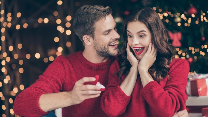 Photo of couple in decorated garland lights room guy giving lady unexpected, engage ring box waiting answer sitting cozy near x-mas tree indoors wear red sweaters