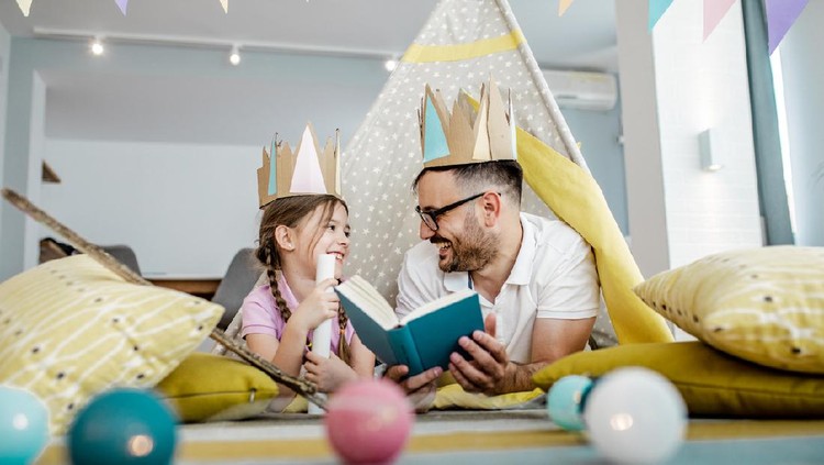 Father and his little girl are on the floor surrounded by toys, he is reading a story to his girl
