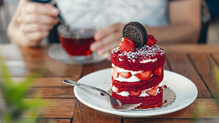red velvet cake with strawberry slice and chokolate cookie and red cup of tea on the brown wooden table in a cafe summer veranda. ands holding a spoon.  Image is  close up