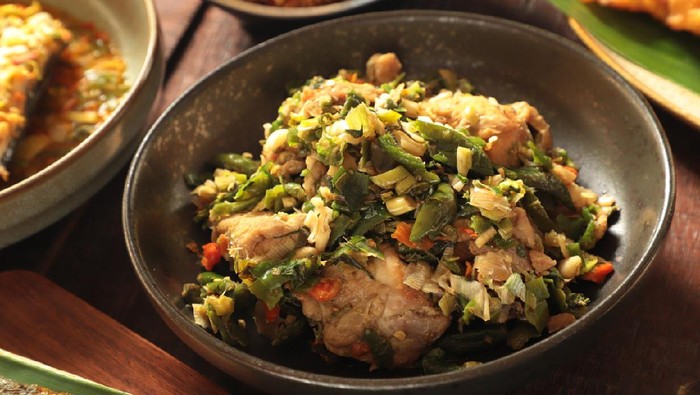 Ayam Woku, a traditional Minahasan dish of braised chicken chunks with lightly-crushed green chili pepper. The dish is plated on a ceramic plate and then placed on a wooden table among other side dishes.