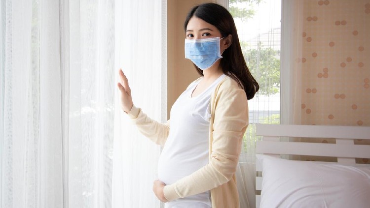 Young beautiful pregnant asian woman standing and touching belly near the window in bedroom. Pregnancy, motherhood wearing virus protection mask mouth concept.