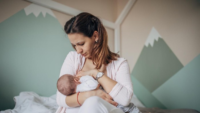 Mother breastfeeding her baby boy in arms, sitting on bed.