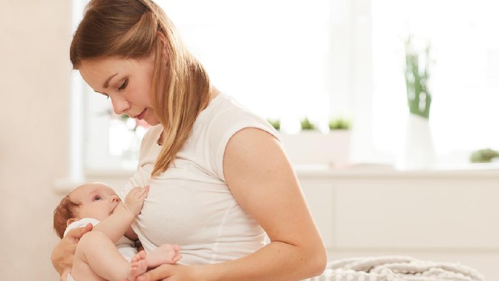 Mother breastfeeding her baby boy in arms, sitting on bed.