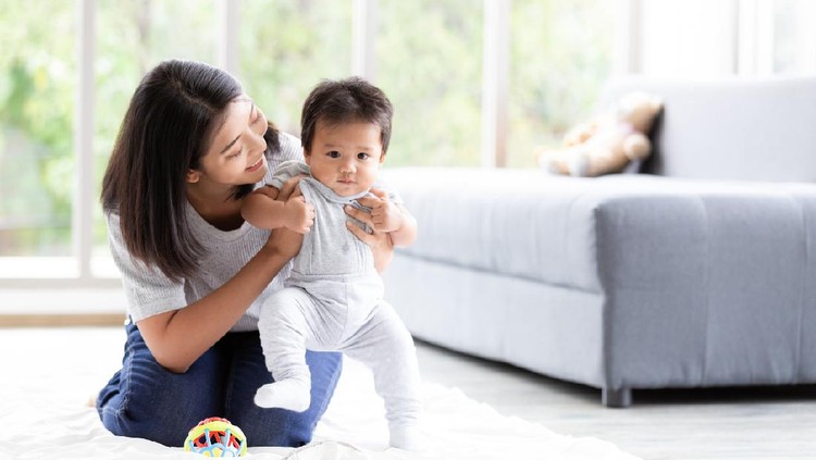 Happy little baby learning to walk with mother help in living room. Baby taking his first steps with mother's help and support with love.