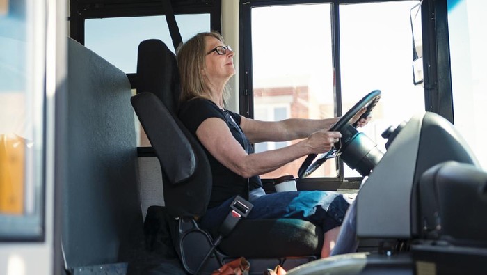 Portrait of mature woman driving a school bus. She ha her security belt on and two hands on the wheel. Horizontal waist up outdoors shot on a bright sunny day. Copy space.