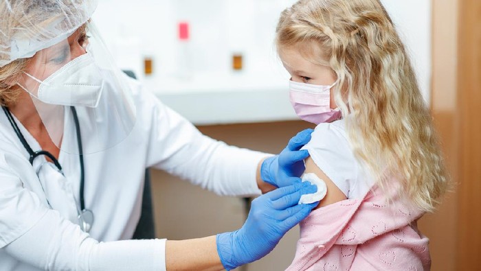 Female doctor in full protective workwear cleaning girl's arm before injecting COVID-19 vaccine, girl wearing protective face mask