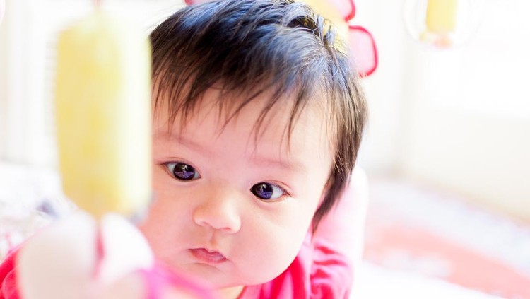 A cute Asian/Eurasian baby girl is fixated on a hanging mobile toy. Her eyes are slightly crossed as she examines it. She is just over 1 years old.