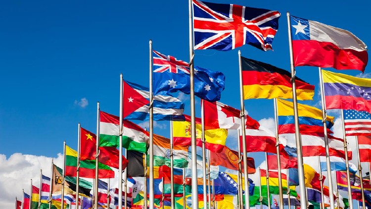 Group of flags of many different nations against blue sky and infront of a convention center.