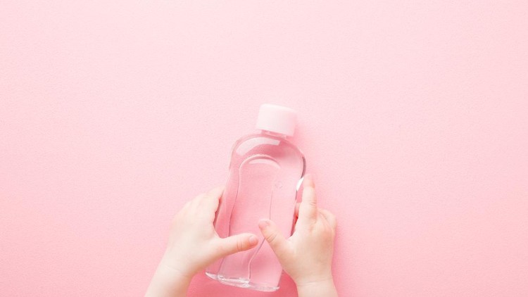 Baby girl little hands holding transparent plastic bottle of oil on light pink table background. Pastel color. Care about soft body skin. Closeup. Point of view shot. Top down view.