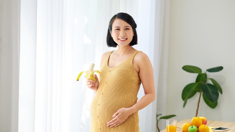 pregnant woman on living room with healthy fruit juice and eating banana