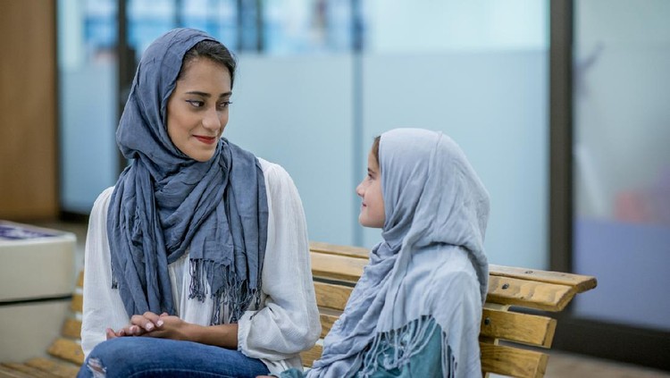 dongeng A Muslim mother and daughter are indoors in a shopping mall. They are both wearing casual clothing and head scarves. They are smiling at each other while sitting on a bench.