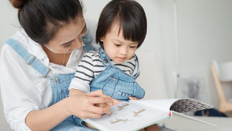 Vietnamese mother and little daughter girl reading a book in the morning together in the living room at home