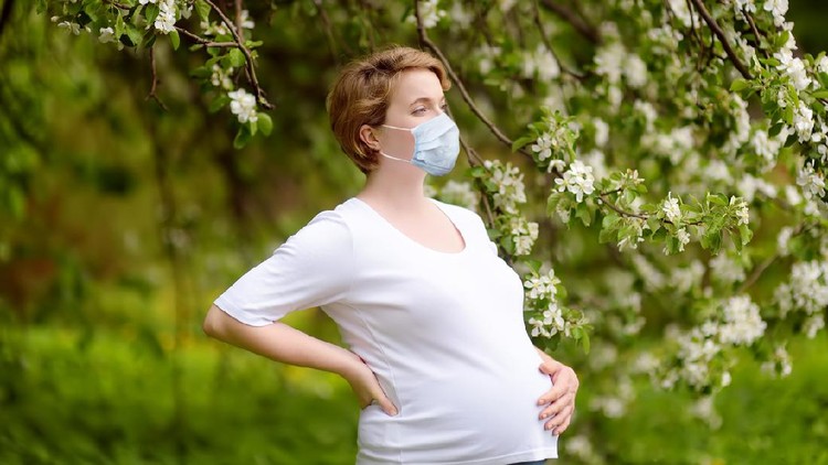 Portrait of a beautiful pregnant young woman wearing disposable medical face mask in the spring park during coronavirus outbreak. Safety in a public place while epidemic of covid-19.