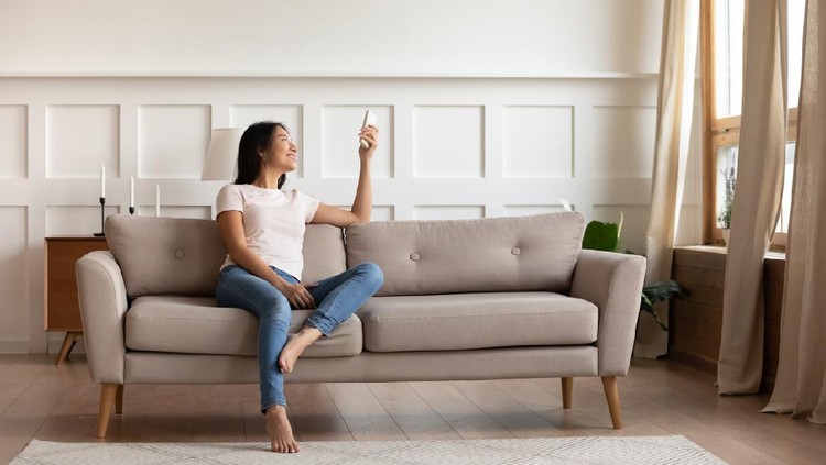 Young asian woman switching on air conditioner while resting seated on couch in modern interior living room, holds remote climate control cooler system manage temperature at contemporary home concept