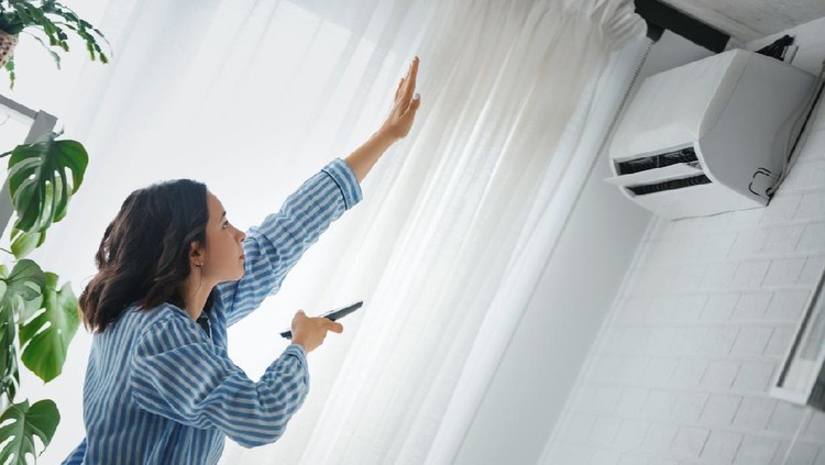 Young asian woman switching on air conditioner while resting seated on couch in modern interior living room, holds remote climate control cooler system manage temperature at contemporary home concept
