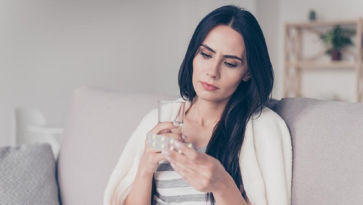 Portrait of troubled ill young woman sitting at home holding glass of water and reading discription of medication in her hand