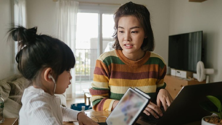 Asian girl doing homework on tablet while young woman is using her laptop at home.