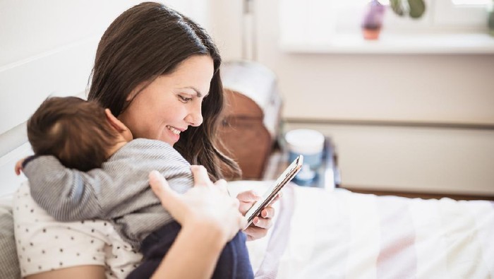 Mother using a mobile phone while breastfeeding her newborn baby boy. They are lying in bed in their bedroom. Belgrade, Serbia