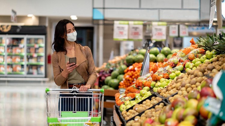 Woman shopping at the grocery store wearing a facemask to avoid the coronavirus while following a list on her cell phone - COVID-19 lifestyle concepts