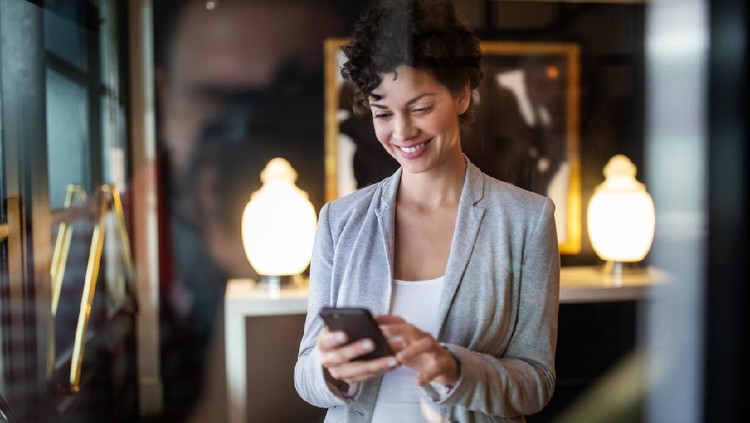 Young businesswoman standing inside a hotel lobby using mobile phone. Female standing at hotel hallway booking online taxi using smart phone app.