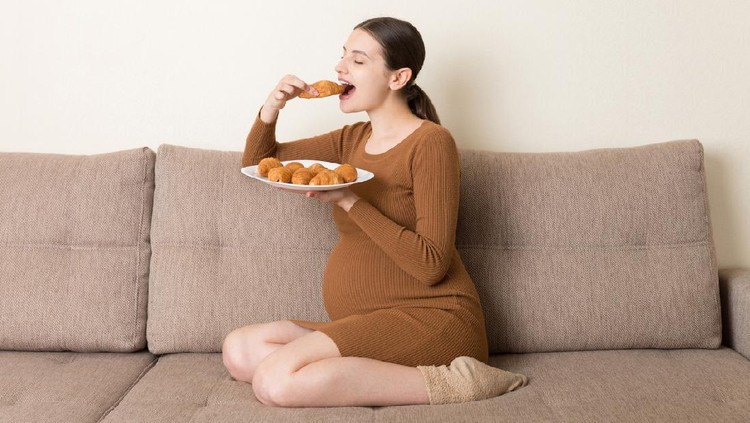 Young pregnant woman enjoys eating croissant on the sofa. Unhealthy pastry during pregnancy concept.
