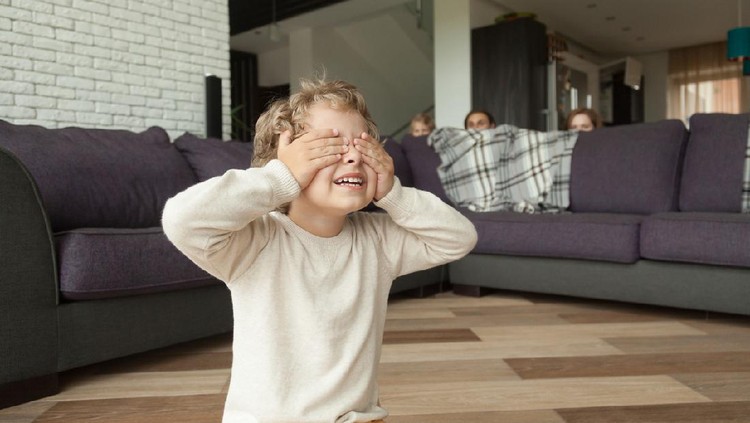 Kid boy playing hide and seek game at home, child closing eyes with hands counting while parents and sister hide behind sofa in living room peeking out, happy family having fun with children concept