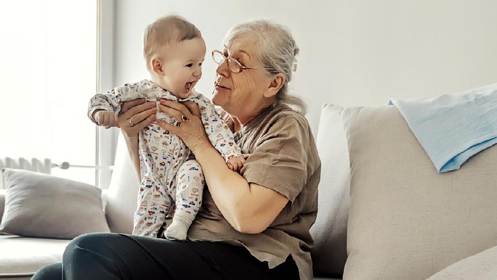 Grandmother holding little grandson in the room at home. Senior woman hold little baby cute smiling. Happy grandmother with her grandchild in home.