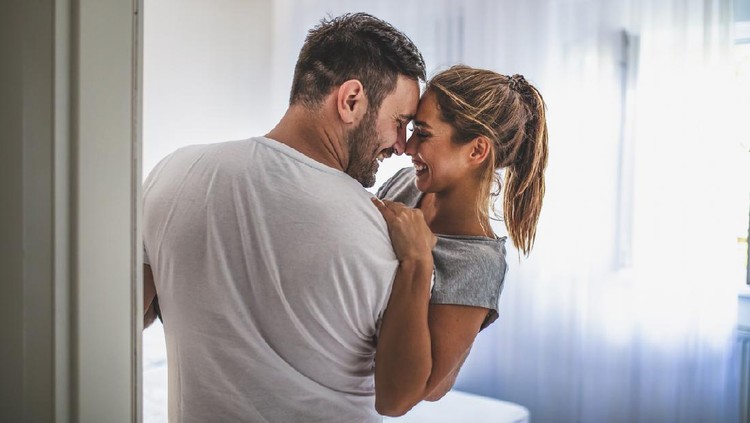 Handsome man in white T-shirt carrying young attractive woman while spending free time at home. Loving young hipster couple relaxing at home and kissing. View from another room through the doorway