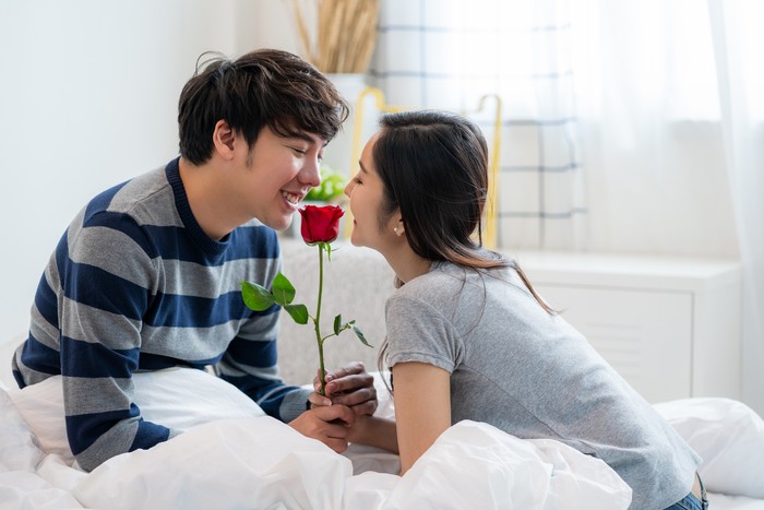 Romantic Asian couple in the bedroom, a man giving a rose to a beautiful woman and both kissing a beautiful rose with love and happiness, Beautiful elegant Asian couple is hugging and smiling in bedroom