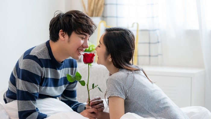 Romantic Asian couple in the bedroom, a man giving a rose to a beautiful woman and both kissing a beautiful rose with love and happiness, Beautiful elegant Asian couple is hugging and smiling in bedroom
