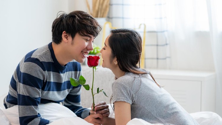 Romantic Asian couple in the bedroom, a man giving a rose to a beautiful woman and both kissing a beautiful rose with love and happiness, Beautiful elegant Asian couple is hugging and smiling in bedroom