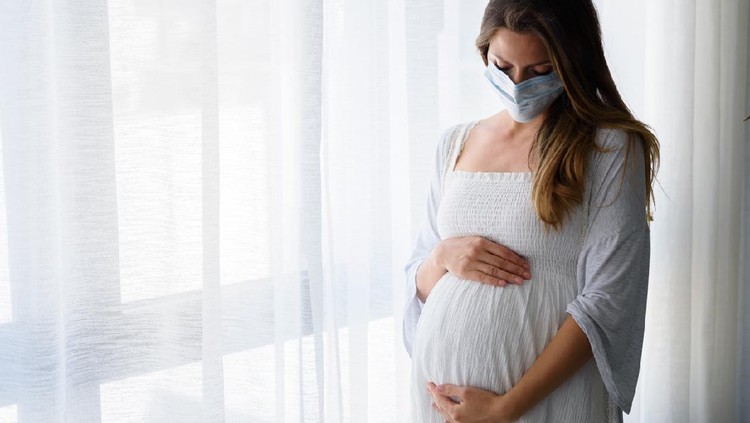 Pregnant woman standing by the big window with face medical mask on. Worries about child birth during pandemic.