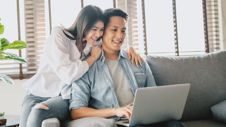 Happy Asian couple using laptop on sofa at home. Woman embracing man from behind