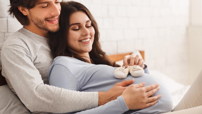 Happy expectants. Smiling pregnant woman holding baby shoes on her belly while husband cuddling her from back on bed at home, closeup
