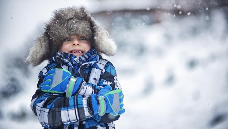 Portrait of a cute boy in big fake fur cap. It's snowing and freezing outside and the little boy is haking the winter day. Snow flakes are falling on the cap.