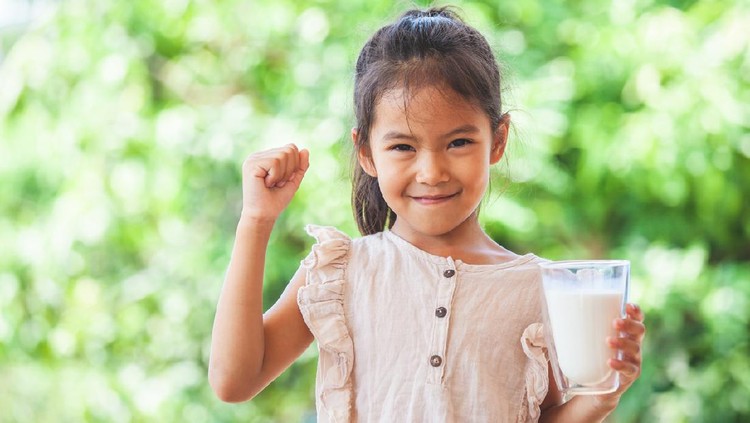 Cute asian child girl holding glass of milk and make strong gesture on green nature background