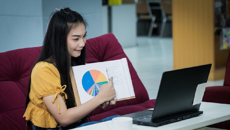 Ilustrasi PJJ A young Asian female university student presents chart on her project study online via laptop at home during COVID-19 pandemic to practice social distancing. Stock photo.