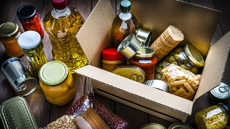 High angle view of a cardboard box filled with multicolored non-perishable canned goods, conserves, sauces and oils shot on wooden table. The composition includes cooking oil bottle, pasta, crackers, preserves and tins. High resolution 42Mp studio digital capture taken with SONY A7rII and Zeiss Batis 40mm F2.0 CF lens