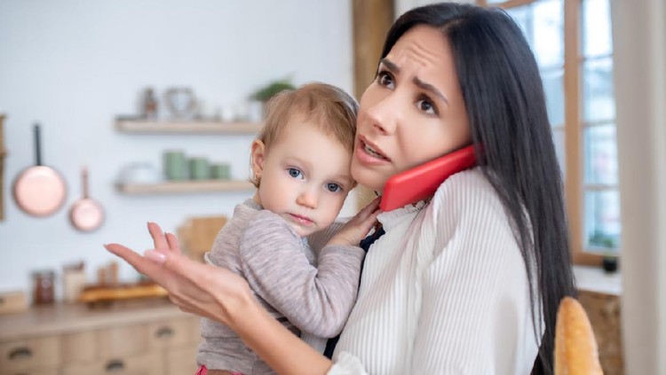 At home. Working mom holding baby, talking on the phone, feeling distressed and worried