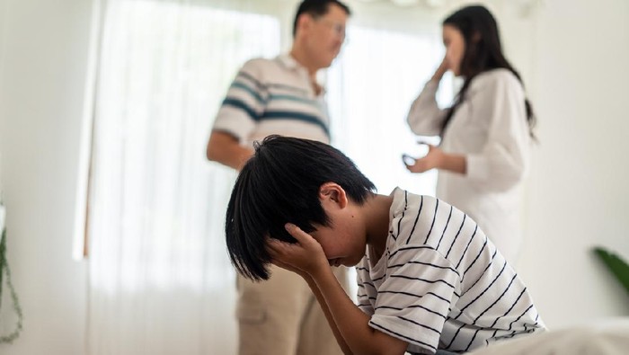 Asian boy kid sitting and crying on bed while parents having fighting or quarrel conflict at home. Child covering face and eyes with hands do not want to see the violence. Domestic problem in family.