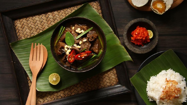 Rawon, the the popular and unique beef soup from East Java. Beef cubes cooked in rich broth flavored with native keluak fruit (pangium edule) and other herbs and spices. Topped with bean sprouts and chives. Accompanied with salted duck egg, red chili paste and steamed rice. The soup is served in a black ceramic bowl, then placed on a woven tray lined with banana leaf. A pair or wooden cutlery and lime half is also placed on the the tray. The accompanying side dishes and condiment are placed near the tray. The meal is arranged on a dark-colored wooden table.