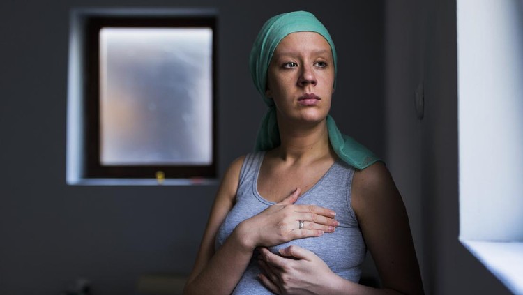 Health, medicine concept - woman in headscarf, fighting breast cancer, checking breast for signs of cancer. Breast examination by home method.  Pensive female examining breast at home while standing next to the window during the day.