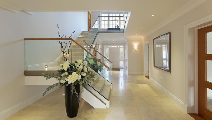 a beautiful entrance hall from a luxury new home with Italian marble flooring and stairs. In the background is a large bright front door area whilst the stairs lead up to the first floor landing with a large Georgian window creating more light. The stairs are sided by wood topped glass banisters. Either side of the stairwell are two large vases with silk flowers.