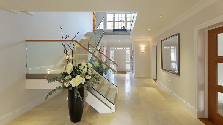 a beautiful entrance hall from a luxury new home with Italian marble flooring and stairs. In the background is a large bright front door area whilst the stairs lead up to the first floor landing with a large Georgian window creating more light. The stairs are sided by wood topped glass banisters. Either side of the stairwell are two large vases with silk flowers.