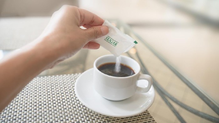 human hand with white sugar packet and pouring in hot black coffee , in soft focus